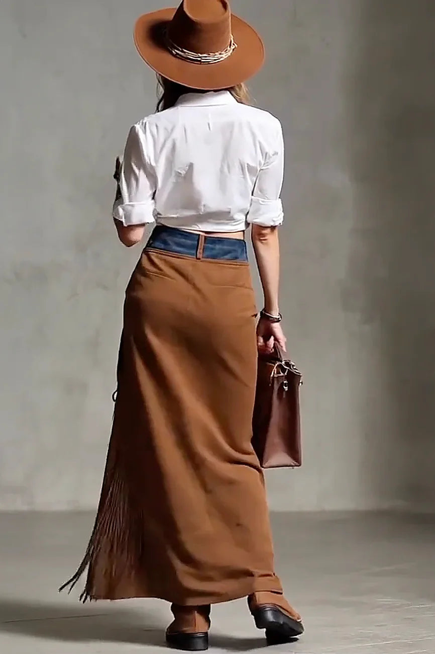 Person wearing a white shirt, brown skirt with fringe, and brown hat against a plain background