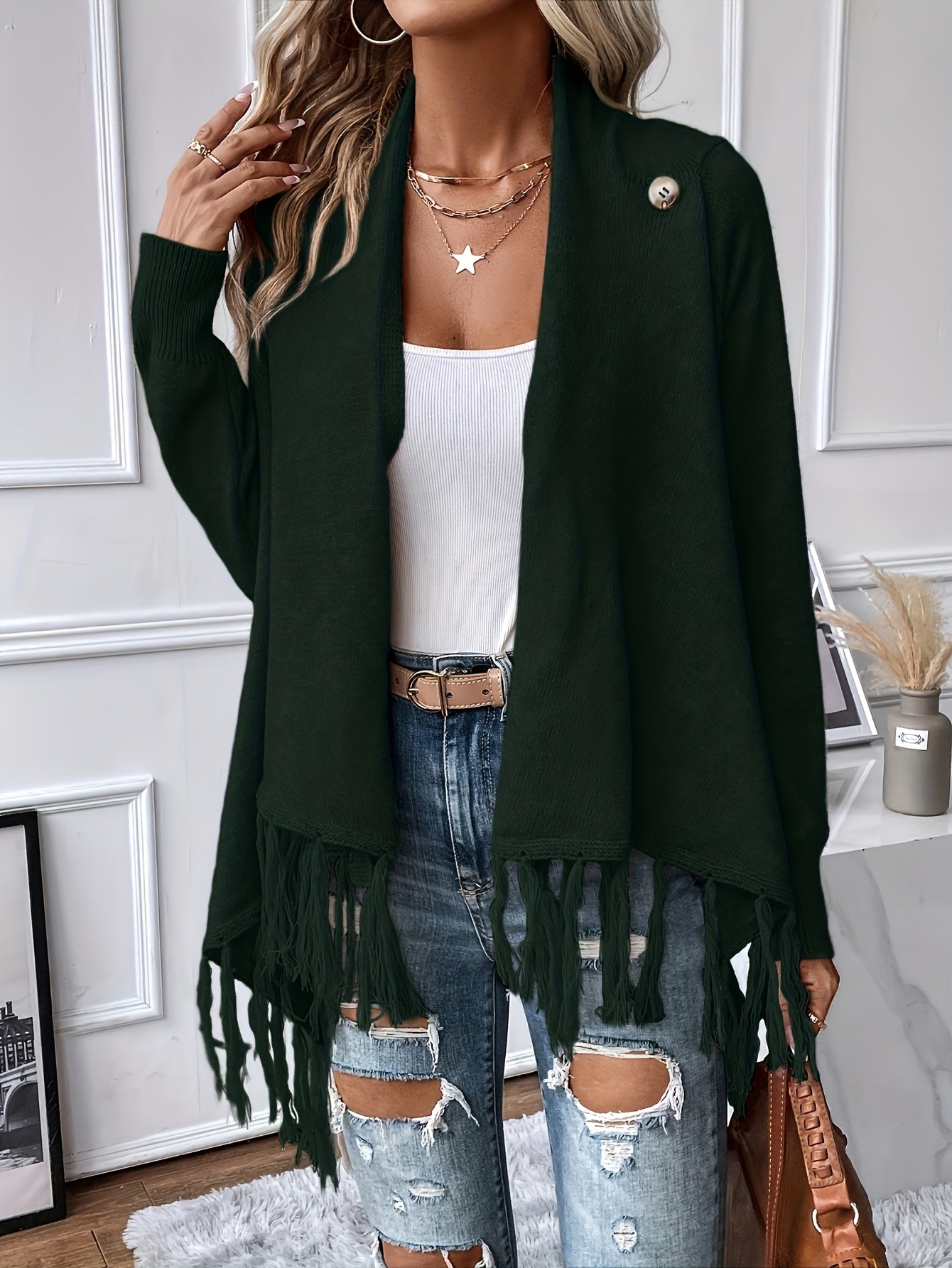Person wearing a dark green cardigan with fringe, white top, and ripped jeans indoors.