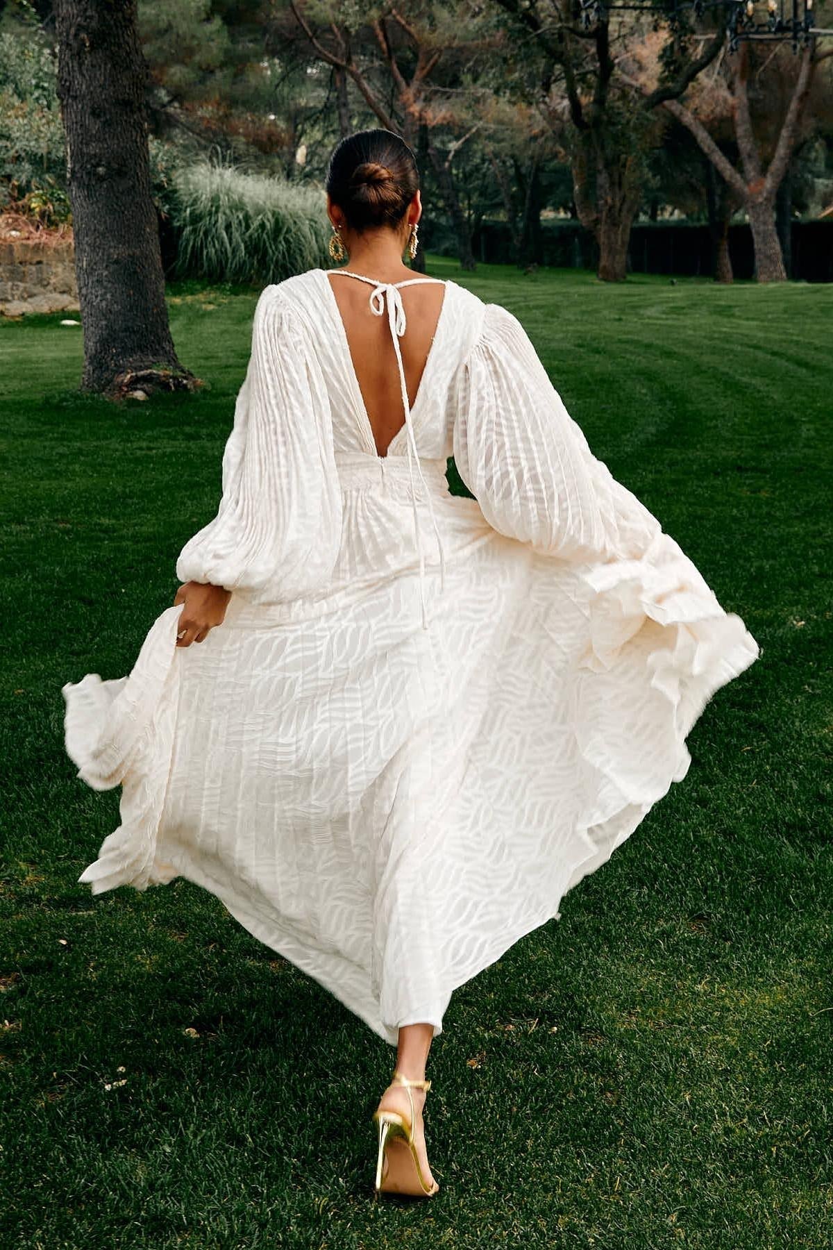 Woman in a white dress with long sleeves and open back walking on grass.