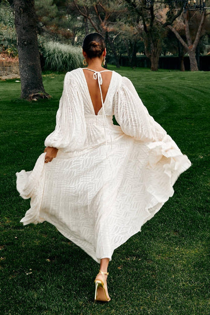 Woman in a white dress with long sleeves and open back walking on grass.